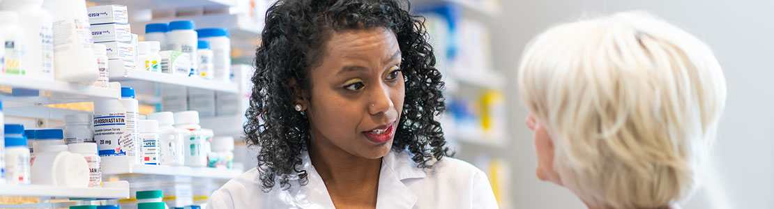 Female pharmacist talking with senior female while standing in front of shelves of pill bottles. 