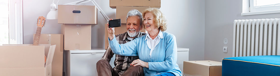 Senior couple smiling as they take a selfie among moving boxes in their home.